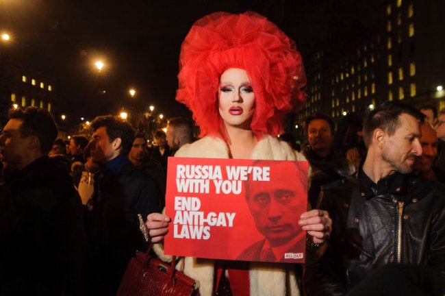 Drag protesta contra as leis anti_gays russa, segurando uma plava vermelha com a foto de Putin. Ela usa uma peruca vermelha e um casaco bege