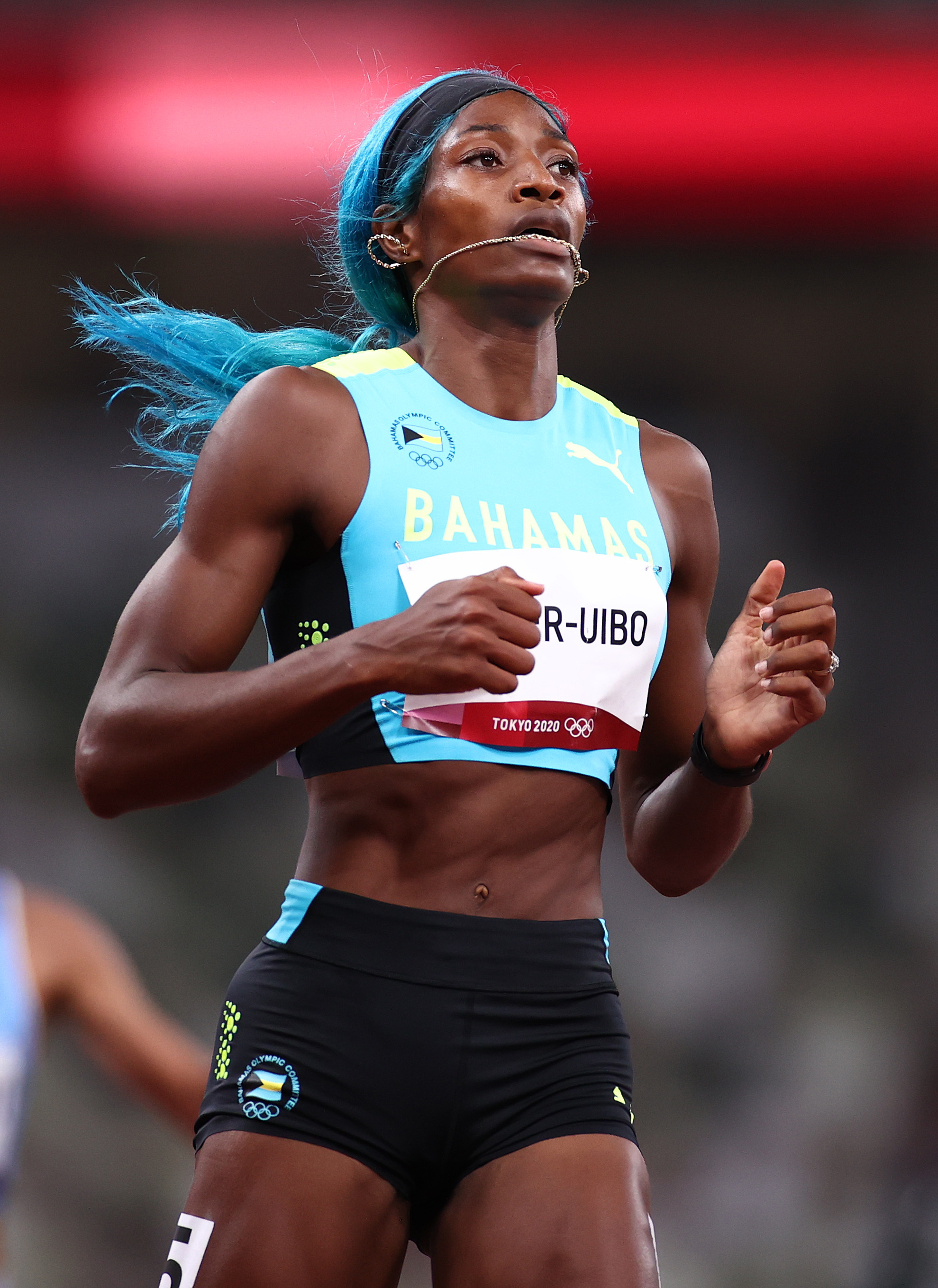 Shaunae Miller-Uibo, das Bahamas, na pista de atletismo das Olimpíadas de Tóquio 2020. Ela está correndo, usando uma lace azul e com um colar na boca.