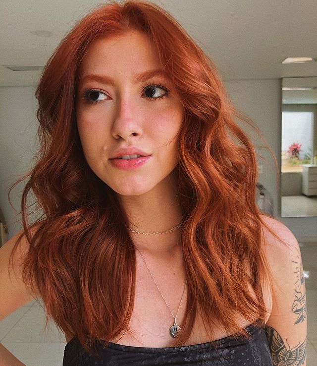 Selfie de uma mulher. Ela usa um top cropped preto, cabelo médio ruivo acobreado ondulado com babyliss e maquiagem natural. Ela olha para o lado e não sorri para a foto.