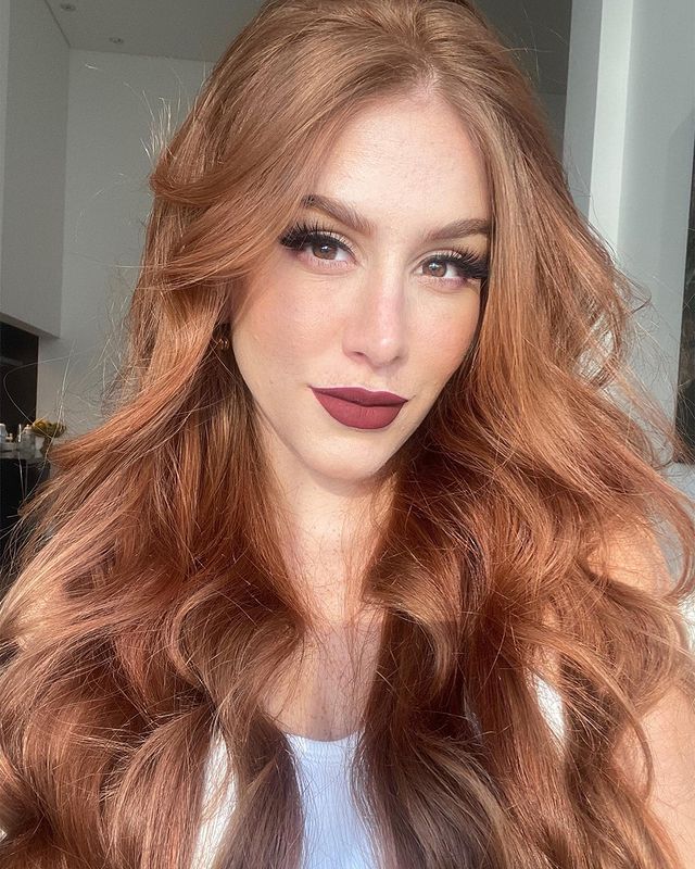 Selfie de uma mulher. Ela usa uma regata branca, cabelo ruivo claro ondulado com babyliss solto e maquiagem com cílios postiços e batom escuro. Ela olha para a câmera e não sorri.