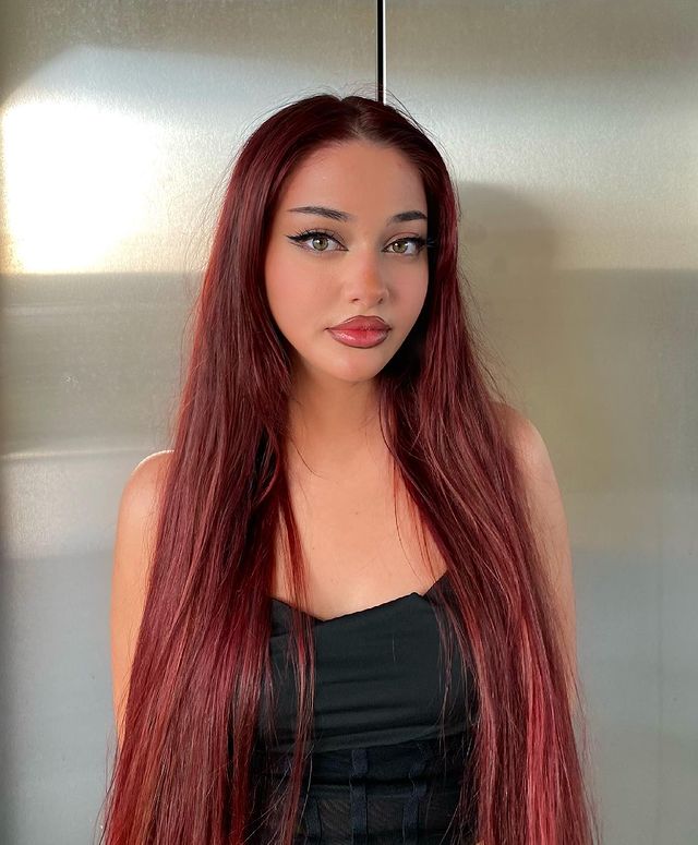 Foto de uma mulher. Ela usa um corset preto, cabelo liso longo e vermelho solto, maquiagem com delineado e gloss nos lábios. Ela olha para a câmera e sorri levemente.