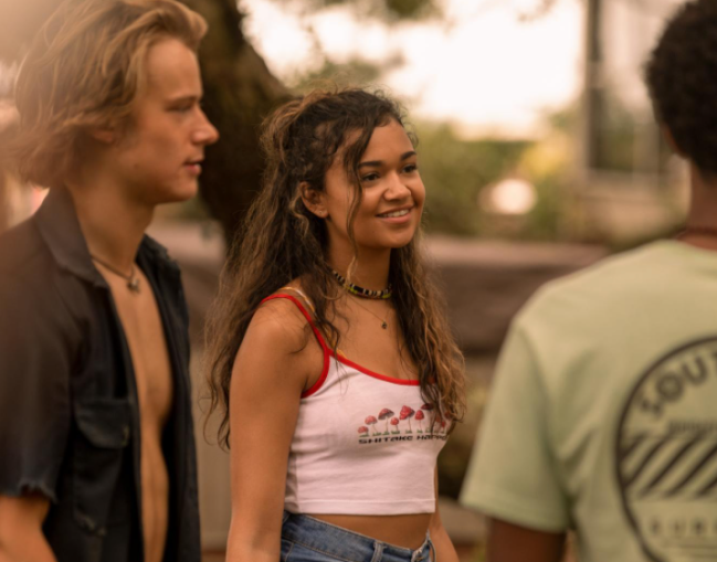 Três personagens de Outer Banks conversando, um rapaz usa camiseta escura aberta, e menina usa cropped branco e short jeans e o último menino usa camiseta verde, ele está de costas.