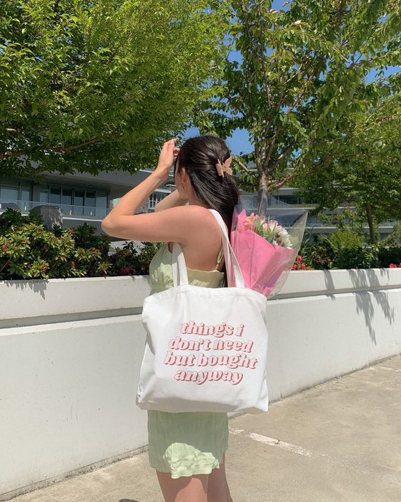 Foto de uma mulher na rua. Ela usa um vestido verde menta, ecobag com estampa rosa claro com um buquê de flores dentro e cabelo preso com uma piranha. Ela está de lado e com a mão esquerda no cabelo, não olha para a câmera e não sorri.