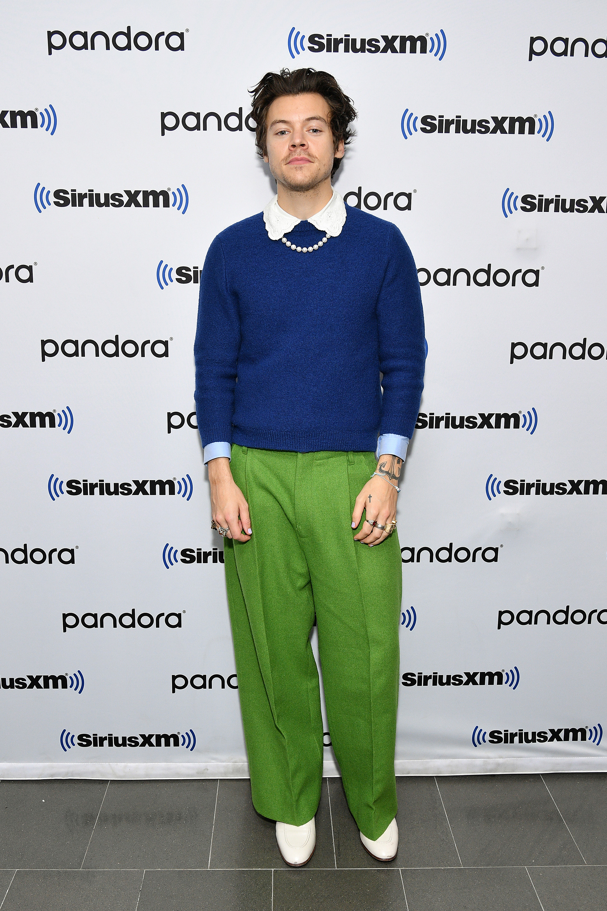 Harry Styles usando look com suéter azul e camisa branca com a gola aparecendo, calça pantalona verde e bota branca, além de colar de pérolas. Ele está olhando para a câmera com expressão facial séria.