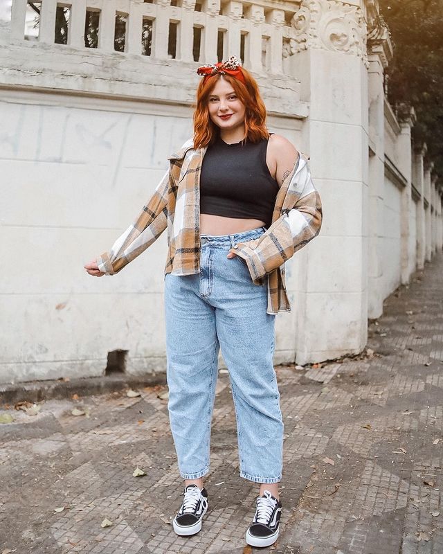 Foto de uma mulher em uma calçada. Ela usa uma regata cropped preta, casaco xadrez em tons de marrom, calça jeans, tênis preto e bandana no cabelo. Ela olha para a foto e sorri levemente.