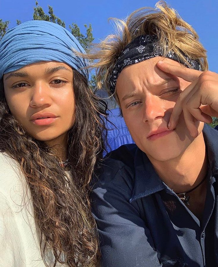 Madison Bailey e Rudy Pankow em selfie; ela está usando uma camiseta branca e lenço azul na cabeça com a expressão séria e ele está de bandana preta com detalhes brancos e uma camisa azul com a mão no rosto e os olhos cerrados por conta do sol que bate no rosto dos dois