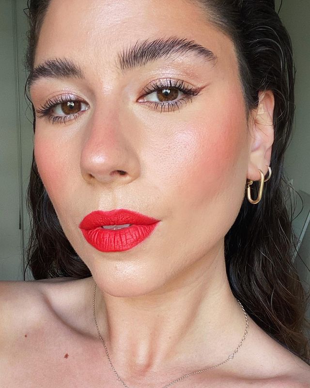 mini-delineado Selfie do rosto de uma mulher. Ela está com o cabelo solto, sobrancelha penteada para cima, pele natural com blush marcado, sombra cintilante, mini delineado gatinho e batom vermelho. Ela olha para a câmera e não sorri.