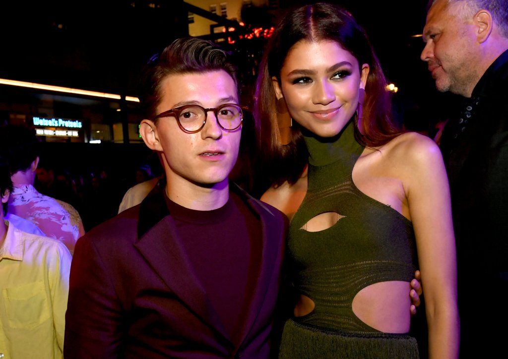 Tom Holland e Zendaya posando para foto na premiere de Homem-Aranha 2; ele está com terno e blusa vinho enquanto Zendaya usa um vestido verde com recortes; ela olha para o lado sorrindo levemente e ele olha para câmera com o braço ao redor da cintura da atriz