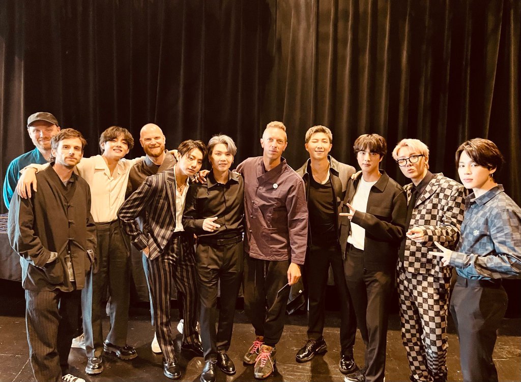 Foto dos integrantes do BTS com a banda Coldplay; eles estão posando para foto abraçados em uma linha horizontal e sorriem para câmera; o fundo é uma cortina escura e a iluminação do ambiente é amarelada