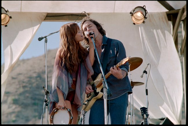 Daisy Jones and The Six; Daisy e Billy cantando no palco e dividindo um microfone enquanto Daisy segura um microfone e Billy toca guitarra