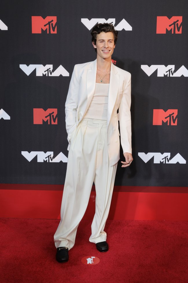 Foto do Shawn Mendes no tapete vermelho do MTV Video Music Awards 2021. Ele usa um terno e calça off white, sapato preto e colar prata. Ele olha para a câmera e sorri.