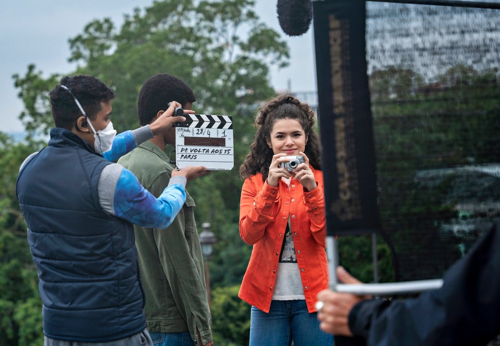 Maisa como Anita nos bastidores de De Volta aos 15, da Netflix, em Paris; ela usa uma calça jeans de lavagem clara com camiseta branca e jaqueta laranja avermelhada e está sorrindo enquanto aponta uma câmera fotográfica pequena para frente e uma claquete com o título do filme está sendo batida pela equipe de filmagens na frente da atriz