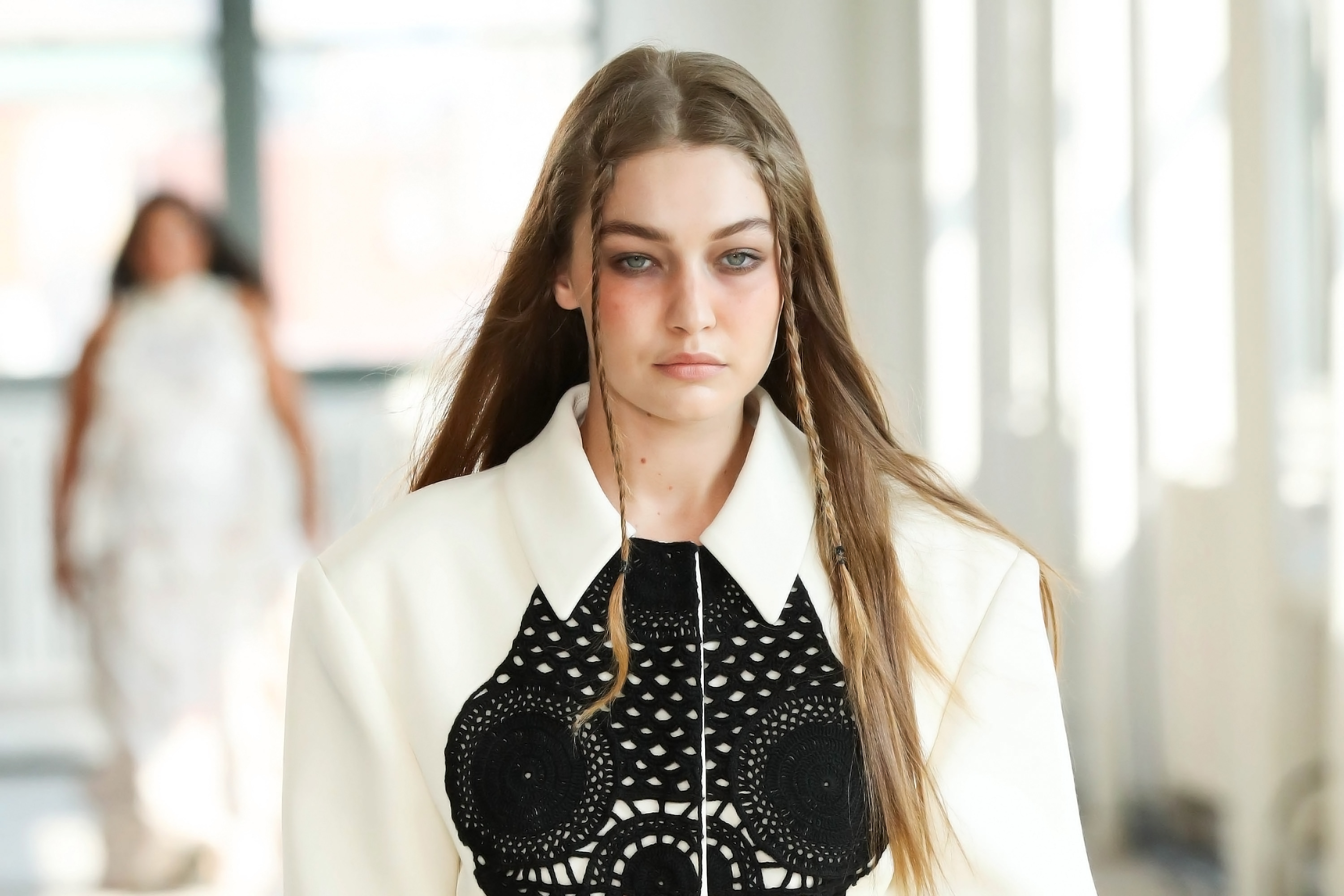 semana-moda-nova-york Gigi Hadid no desfile de primavera-verão 2022 da Altuzarra na semana de moda de Nova York. Ela está com expressão facial séria, trancinhas na franja, camisa branca e detalhe preto no decote.