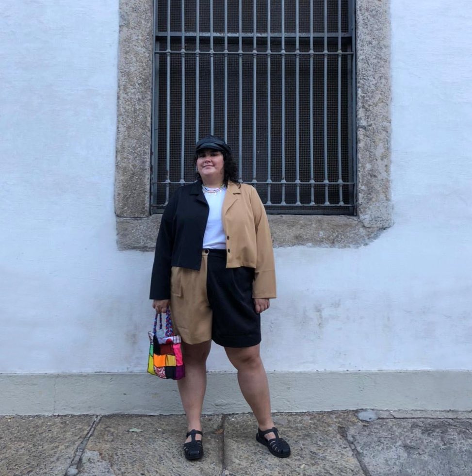 look marrom com sandalia pescador Jovem posando frente a parede branca usando conjunto de peças preta e marrom.