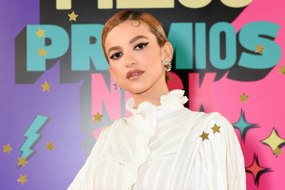 Manu Gavassi nos Meus Prêmios Nick 2021; ela está com posando no painel do orange carpet com expressão séria e usa uma blusa branca de gola alta; estrelas amarelas decoram a imagem