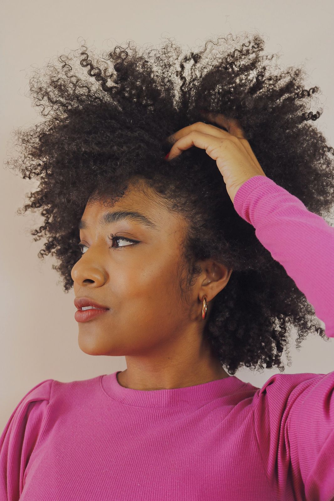 A influenciadora Sah Oliveira usando blusa rosa de manga longa, olhando para o lado com uma das mãos em seu cabelo crespo