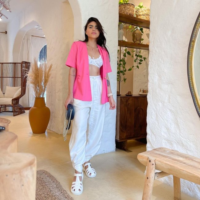 sandalia pescador branca Jovem posando no meio de um cômodo com paredes brancas; ela usa top branco e camisa rosa aberta por cima; calça branca e sandália da mesma cor.