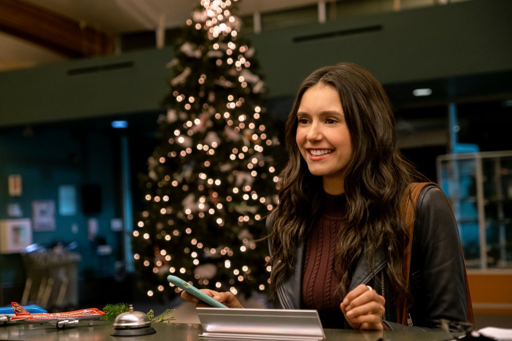 Nina Dobrev como Natalie Bauer em Love Hard, comédia romântica natalina da Netflix; ela está sorrindo e usa uma jaqueta de couro preta com blusa de tricô vinho, a alça da bolsa marrom é aparente no ombro e o cabelo está solto; ela está segurando o celular com capinha azul em uma mão e apoiando a outra no balcão; ao fundo é possível ver uma árvore de Natal com o pisca pisca aceso; está de noite e ela está de frente para um balcão em uma recepção que conta com duas miniaturas de avião, uma azul e outra laranja