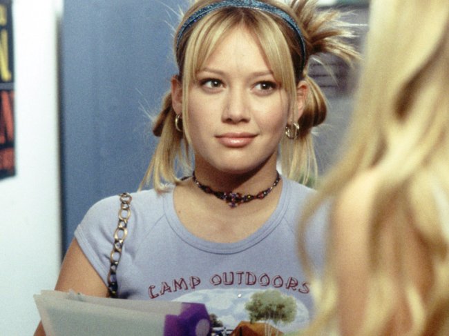 Hillary Duff como Lizzie McGuire com coques nas laterais do cabelo, tiara e blusa cinza.