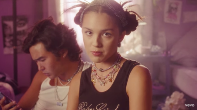 Olivia-Rodrigo-space-buns Print de cena do clipe de Brutal. Podemos ver Olivia Rodrigo com uma expressão séria ao lado de um rapaz.