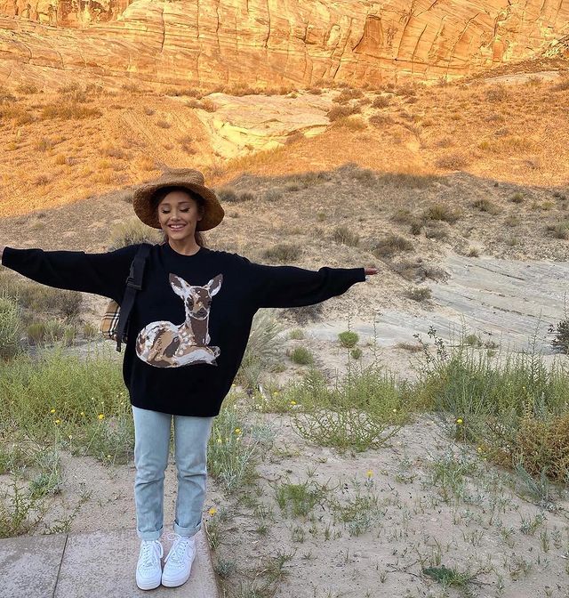 ariana-grande-calca-jeans Foto da Ariana Grande em uma paisagem. Ela usa um chapéu marrom, moletom preto, calça jeans, tênis branco e mini mochila. Ela está sorrindo de olhos fechados e com os braços abertos.