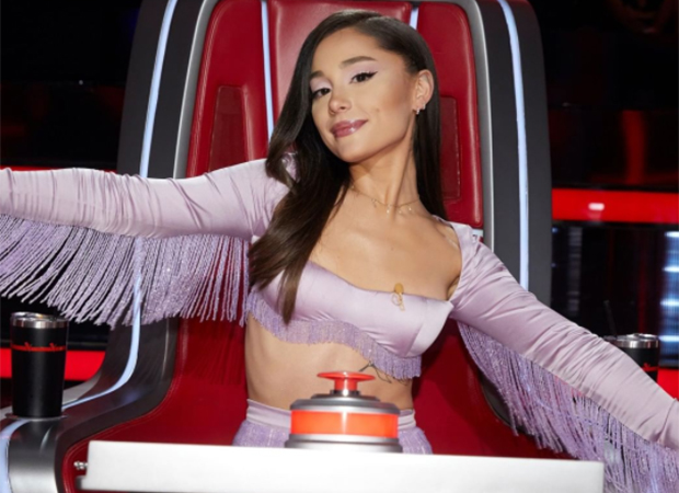 ariana-grande-jurada-the-voice Ariana Grande sentada na cadeira de jurado do The Voice. Ela usa top roxo com franja nas mangas e está com os braços abertos e um leve sorriso no rosto