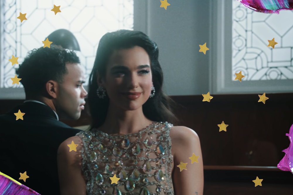 Dua Lipa no clipe de We're Good; ela está sorrindo levemente com um vestido de pedraria prata sem mangas com brincos também prata; ela está sentada de costas para um piano marrom e é possível ver um homem de terno preto tocando o instrumento no segundo plano; no terceiro plano as paredes são brancas e o ambiente é iluminado por janelas também brancas que refletem a luz do dia; manchas de tinta nos tons de lilás, roxo, azul e amarelo decoram as bordas da imagem assim como estrelas amarelas que são usadas como decoração