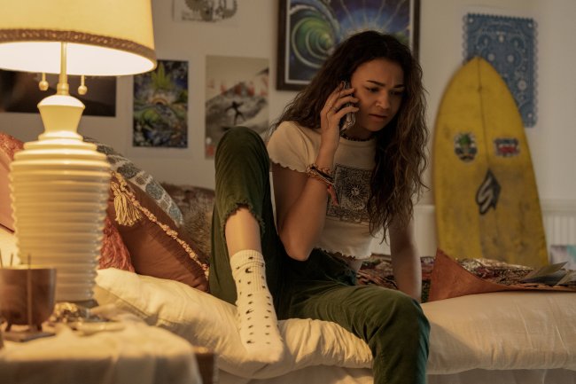 Madison Bailey como Kiara em cena de Outer Banks; ela está sentada na cama conversando no celular usando uma calça verde com blusa branca e meia branca com detalhes; a parede branca está repleta de quadros e uma prancha amarela está encostada no fundo; um abajur ilumina o cômodo com luz amarela