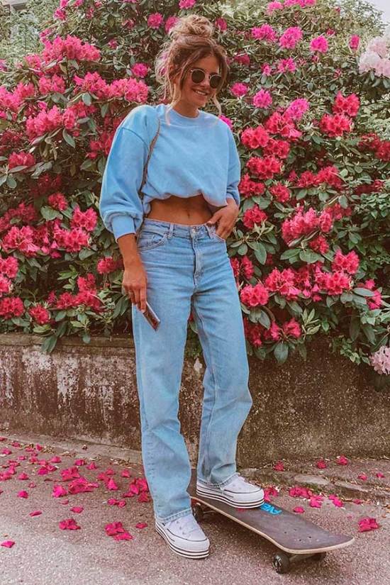 Jovem posando em frente a parede com flores cor de rosa. Ela posiciona a mão direita no bolso e usa calça jeans.