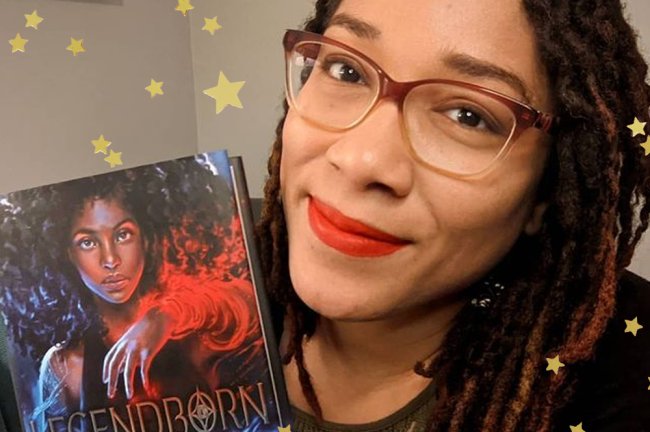 Selfie de Tracy Deonn, autora do livro Lendários, segurando a versão da obra em inglês; ela sorri levemente e está usando óculos e batom vermelho; estrelas amarelas decoram a imagem