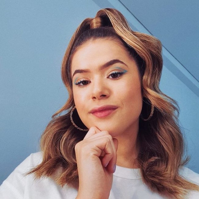 Penteado meio preso Maisa Maisa com penteado meio preso posando para foto com fundo branco e usando camiseta branca, ela está com uma expressão sorridente.