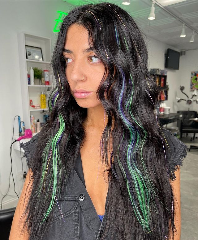 Foto de uma mulher em um salão de beleza. Ela usa uma camisa preta e cabelo preto solto e ondulado com pequenas mechas com fios de glitter verdes. Ela olha para o lado e não sorri.