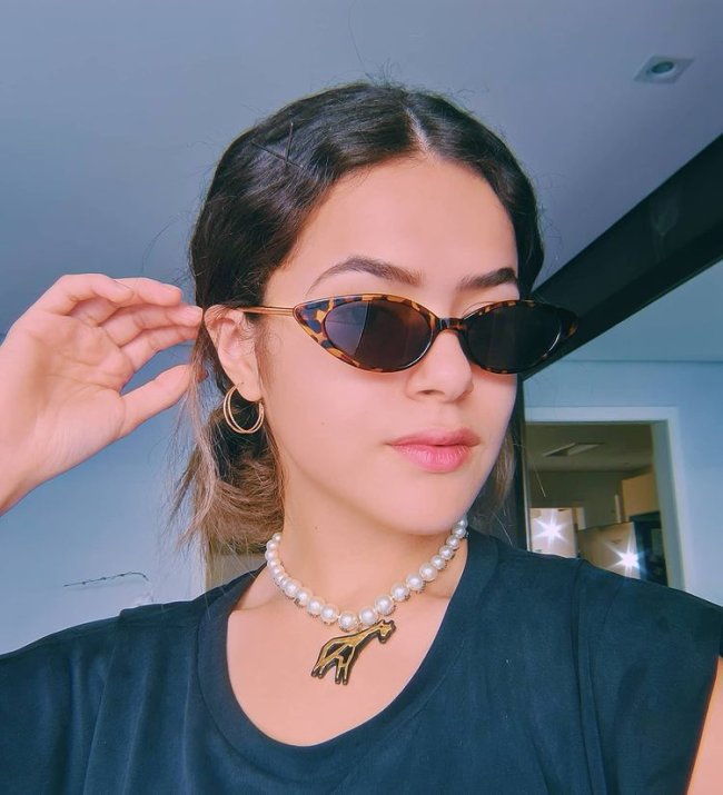 Maisa posando com coque baixo Maisa posando para foto com uma das mãos apoiadas na cabeça, usando óculos escuro e com blusa preta com colar.