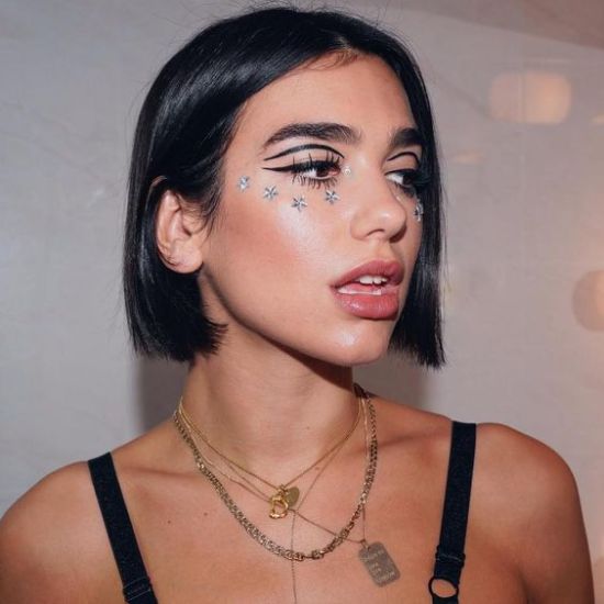 delineado-banana-dua-lipa Foto da cantora Dua Lipa. Ela usa um top preto de alcinha, colares, cabelo solto e maquiagem com delineado preto no formato banana. Ela olha para o lado e não sorri.