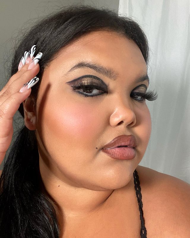 delineado banana Selfie de uma mulher. Ela usa uma blusa preta de alcinha, cabelo preso e maquiagem com delineado banana preto. Ela está com a mão direita no cabelo, olha para a câmera e não sorri.