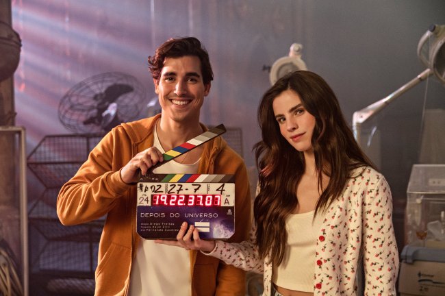 Henry Zaga e Giulia Be nos bastidores de Depois do Universo; os dois estão seguando uma claquete com o nome do filme; Henry sorri e usa uma blusa branca com casaco laranja por cima enquanto Giulia usa uma blusa branca com casaco branco com detalhes de flores vermelhas e sorri levemente