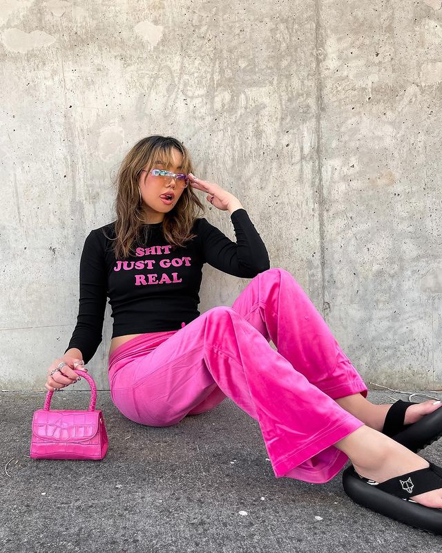 look-com-calca-rosa Foto de uma mulher sentada no chão. Ela usa uma blusa de manga longa preta com detalhes rosa, calça rosa, chinelo preto, bolsa rosa e óculos de sol. Ela está com a mão esquerda no óculos e segura a bolsa com a mão direita, olha para o lado e mostra a língua.