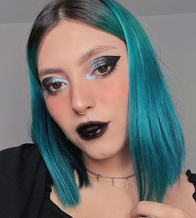 Selfie de uma mulher. Ela usa o cabelo azul e curto solto e liso, maquiagem com delineado azul e preto com batom preto com gloss. Ela olha para a câmera e não sorri.