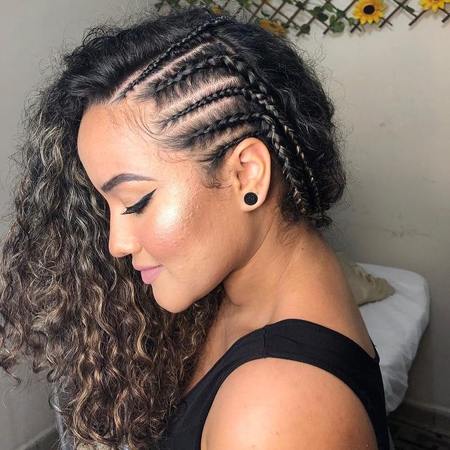 Foto de uma mulher de perfil. Ela usa um blusa preta, maquiagem com delineado gatinho, brinco preto e cabelo cacheado solto com trança na raiz do cabelo. Ela está de perfil, olha para baixo e sorri levemente.