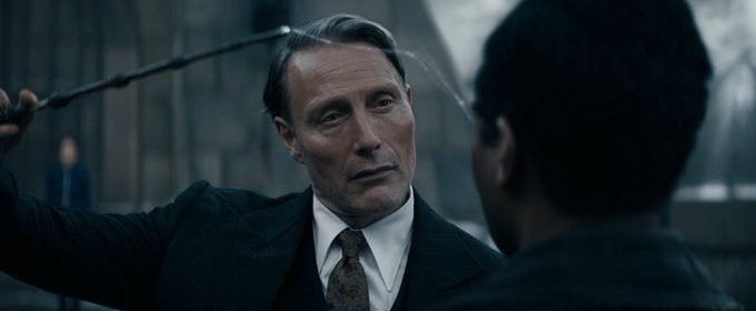 Mads Mikkelsen caracterizado como Gellert Grindelwald; ele está usando terno e gravata na cor preta e camisa branca; a expressão dele é séria e ele segura uma varinha com uma luz branca leve apontada para o rosto de outra pessoa que está de costas para câmera