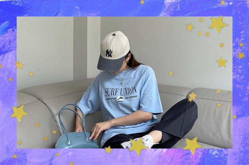 Montagem com o fundo roxo e detalhe de estrelas douradas com a foto de uma mulher. Ela está sentada em um sofá, usa camiseta oversized azul, calça preta, boné, bolsa baguete azul e tênis esportivo branco. Ela olha para baixo e não sorri para a foto.