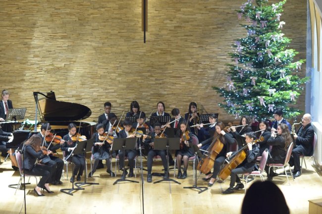 Foto de Marine, da Galera CH, durante intercâmbio. Ela se apresenta com a orquestra do colégio na festa de sinal de ano. Tem uma árvore de Natal enorme atrás dos instrumentistas