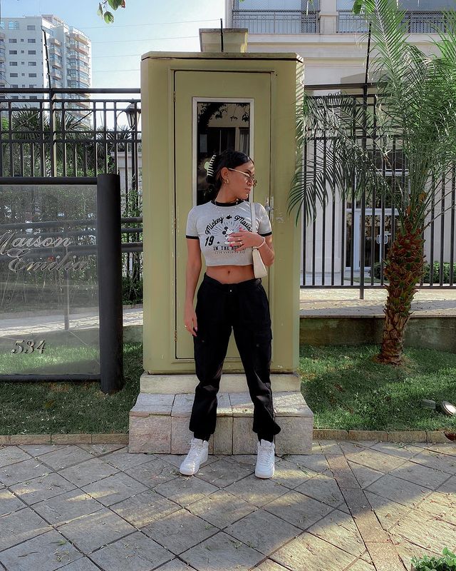 look-calca-baby-tee Foto de uma mulher na rua. Ela usa uma calça baggy preta, baby tee branca e tênis branco. Ela olha para o lado e não sorri.