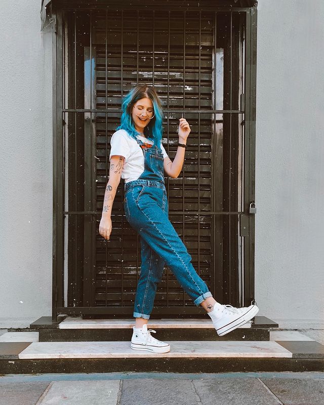 look-com-macacao-jeans Foto de uma mulher em frente a uma porta. Ela usa uma camiseta branca, macacão jeans e tênis branco de cano alto. Ela olha para baixo e sorri.