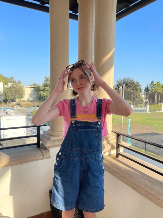 look-com-macacao Foto de uma menina. Ela usa uma camiseta colorida, jardineira jeans e óculos de sol na cabeça. Ela olha para o lado e sorri levemente.