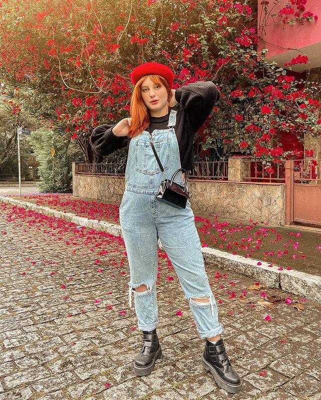 look-macacao-jeans-frio Foto de uma mulher na rua. Ela usa um moletom preto, macacão jeans, coturno preto e bolsa preta. Ela está com as duas mãos no pescoço, olha para a câmera e não sorri.