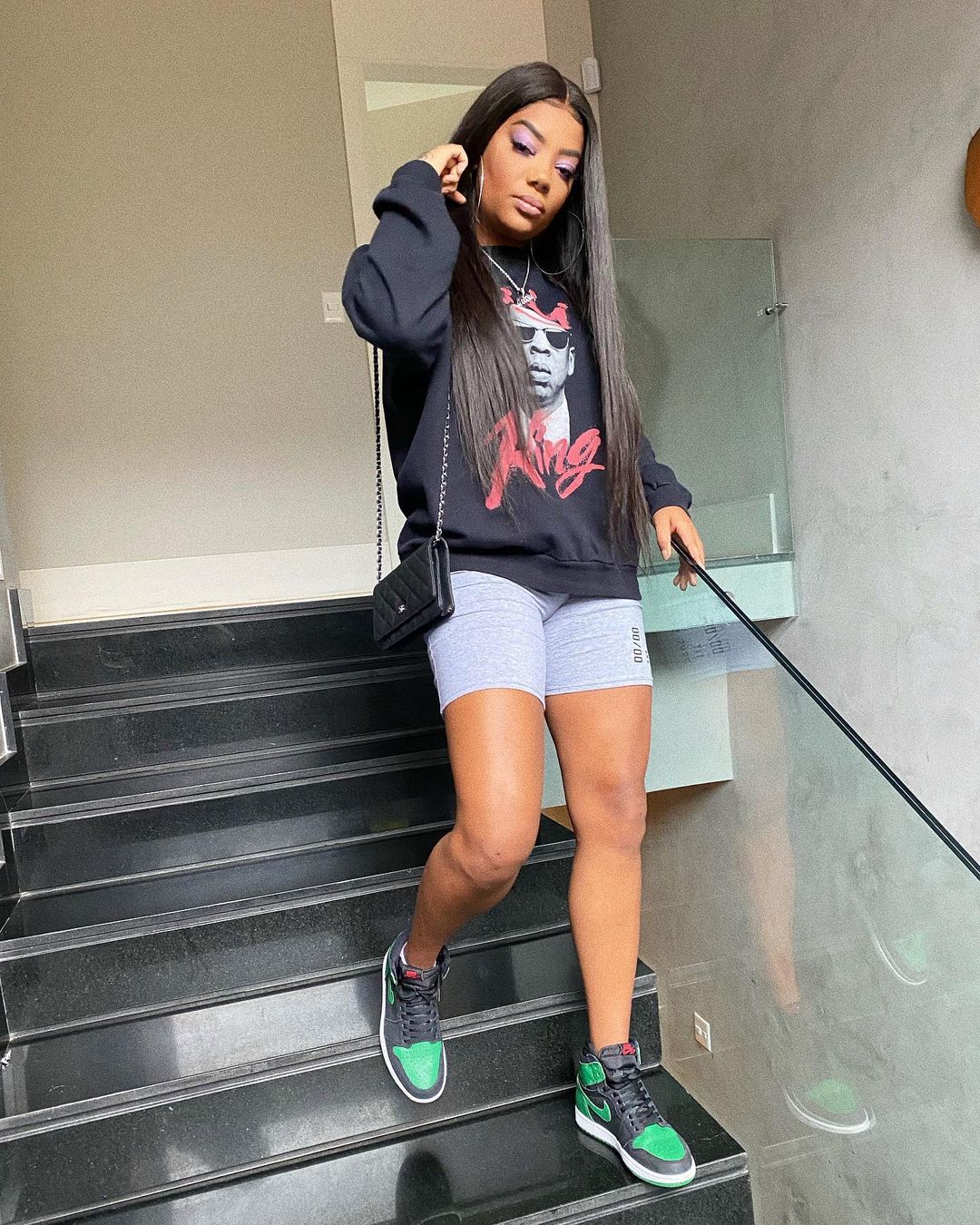 Ludmilla descendo escadas usando look com blusa de moletom preta, biker short cinza, tênis Nike verde e preto e bolsa preta. Ela está com uma das mãos no corrimão e a outra no cabelo.