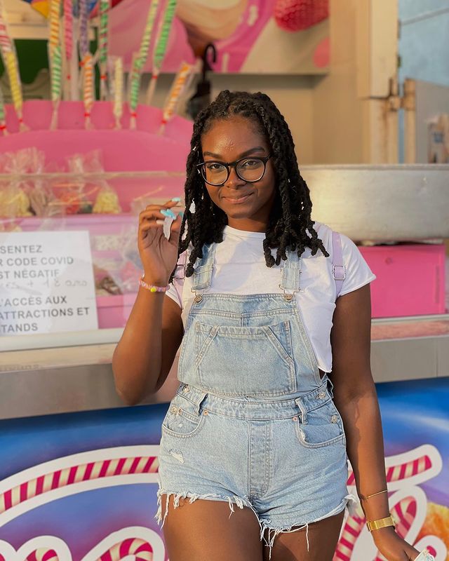 macacao-jeans-curto Foto de uma mulher em frente a uma banca de doces. Ela usa uma camiseta branca e jardineira curta jeans. Ela olha para a câmera e sorri.