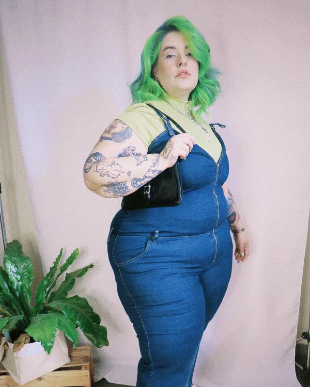 macacao-jeans-look Foto de uma mulher. Ela usa um macacão jeans, camiseta verde e bolsa baguete preta. Ela olha para a câmera e não sorri.