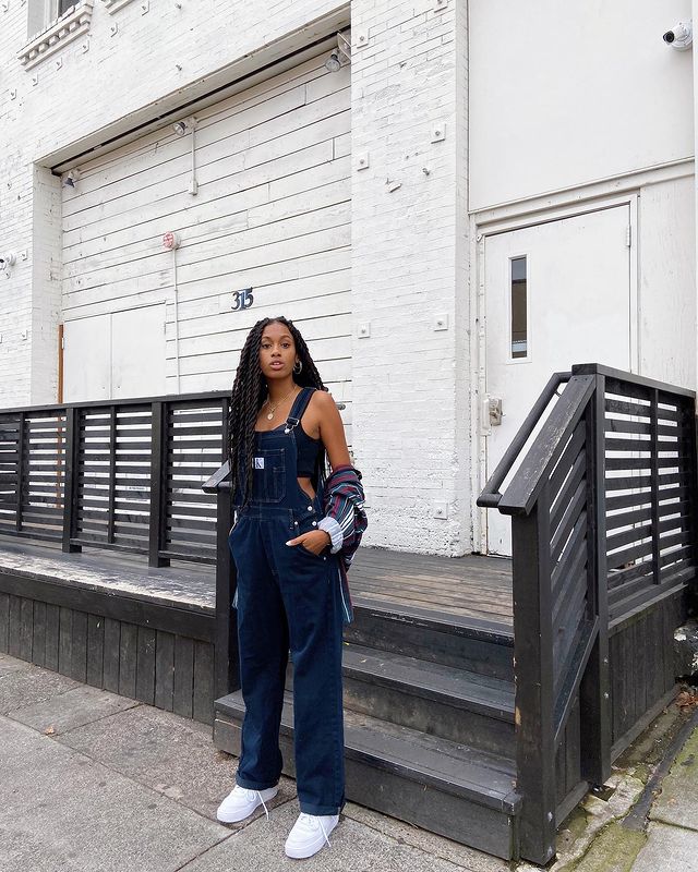 macacao-jeans-look Foto de uma mulher em uma calçada em frente a uma escada. Ela usa um macacão jeans, top cropped preto e segura uma camisa azul listrada. Ela está com as mãos nos bolsos do macacão, olha para a câmera e não sorri.